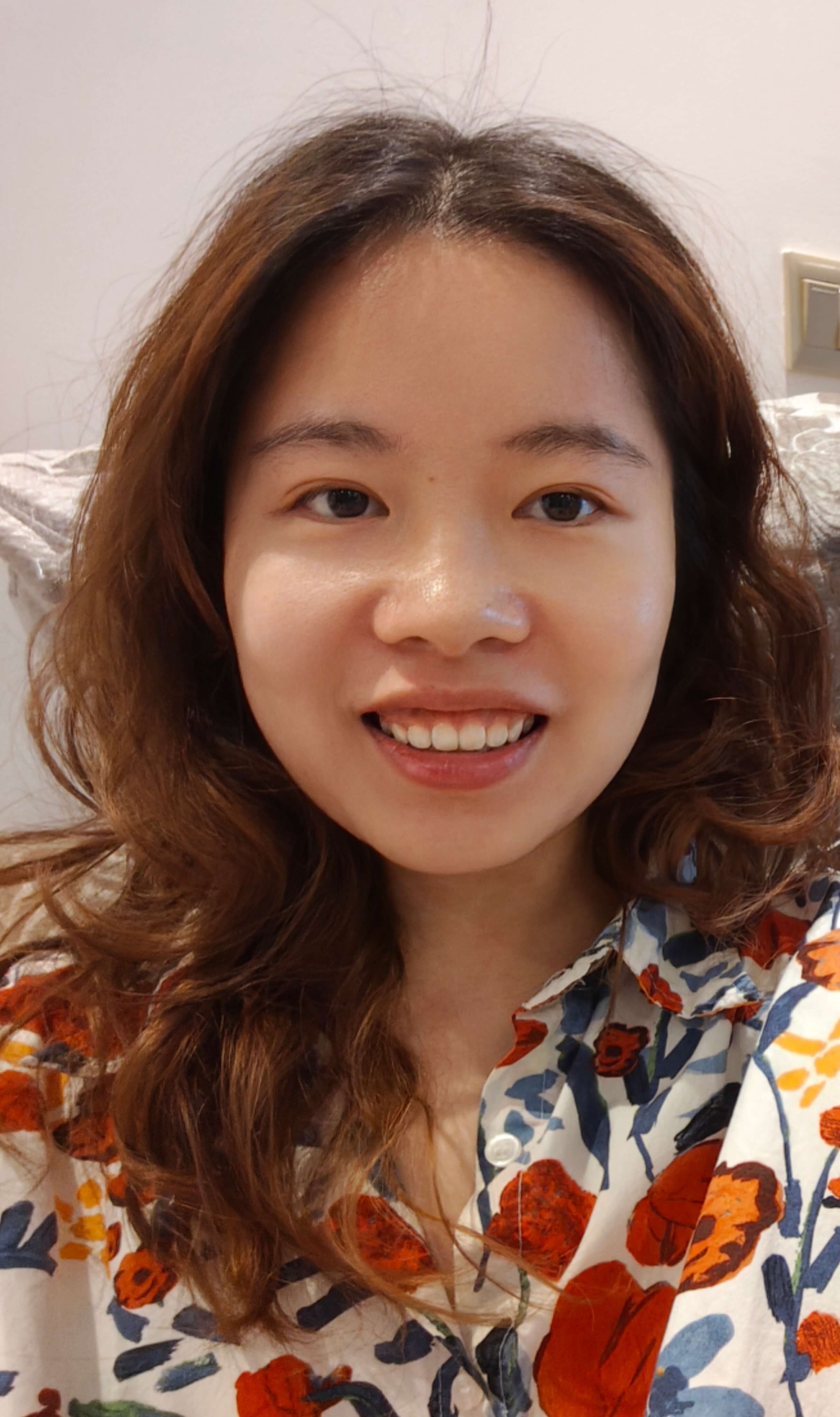 A smiling woman with wavy brown hair, wearing a white shirt with orange and blue floral patterns, in a bright indoor setting.
